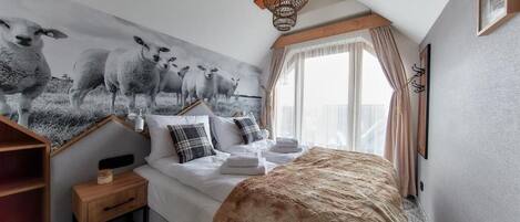 Une chambre à coucher avec un grand lit, une table de chevet, une étagère en bois, un tableau représentant des moutons accroché au mur, un luminaire suspendu et une fenêtre avec des rideaux.