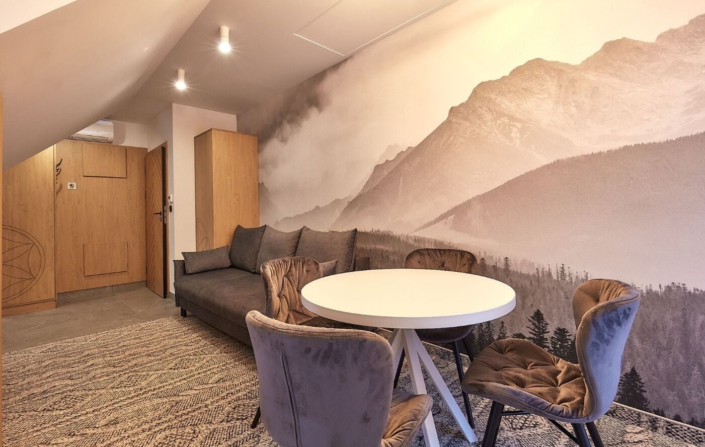 A room with a round table, four chairs, a sofa, and a mural of a mountain landscape on the wall.
