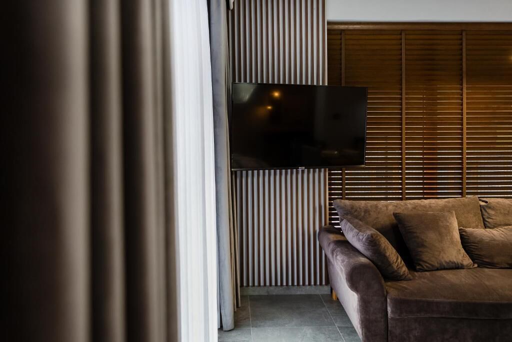 A modern living room with a flat-screen TV mounted on a wooden panel wall, a brown sofa with cushions, and a large window with blinds.