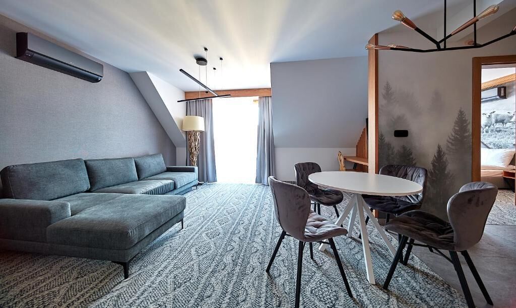 Een moderne woonkamer met een grijze bank, een ronde tafel met stoelen, een kamerbreed tapijt en een groot raam met gordijnen.
