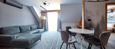 Een moderne woonkamer met een grijze bank, een ronde tafel met stoelen, een kamerbreed tapijt en een groot raam met gordijnen.