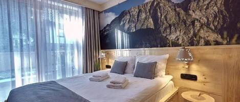 A hotel room with a large bed, a wooden headboard, a nightstand with a lamp, and a mountain mural on the wall.