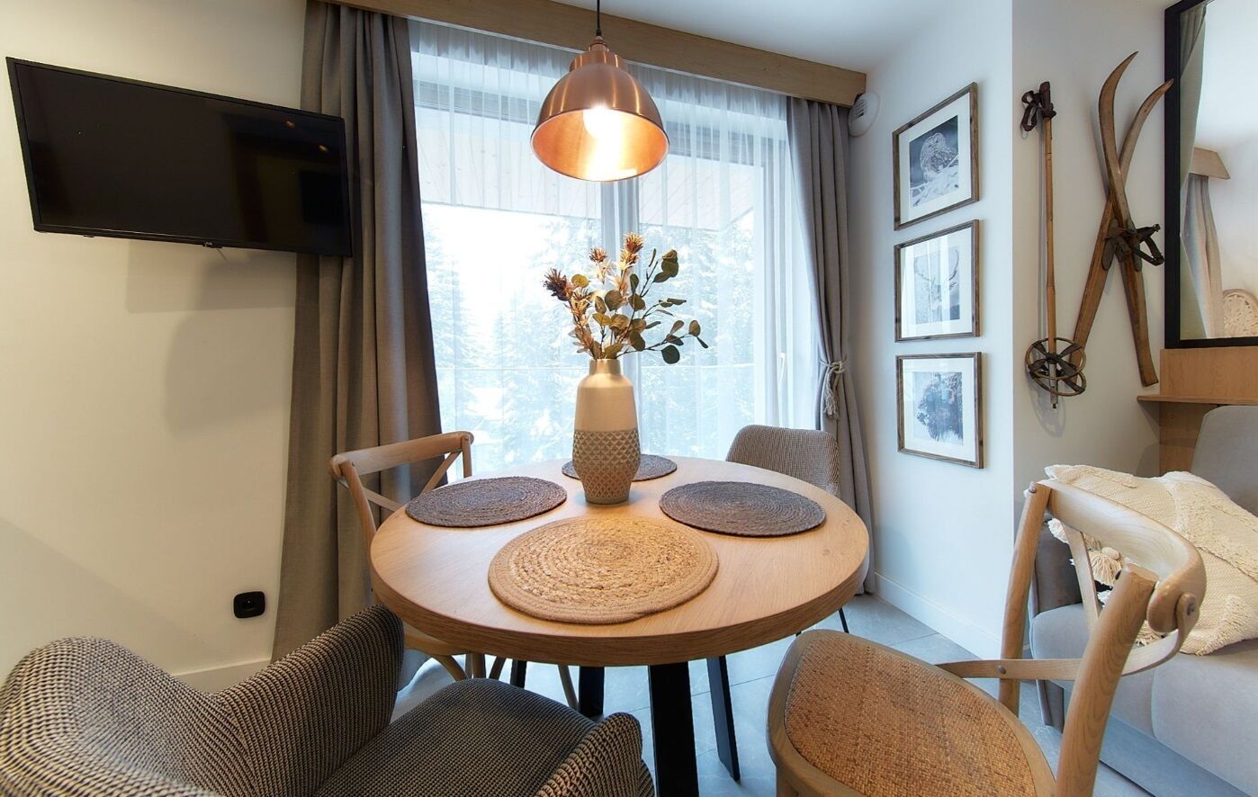A dining area with a round table, four chairs, a vase with dried flowers, a television mounted on the wall, and a window with curtains.