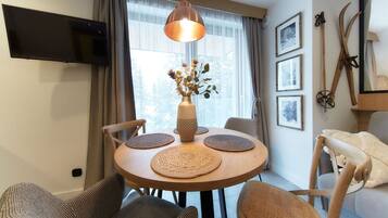 A dining area with a round table, four chairs, a vase with dried flowers, a television mounted on the wall, and a window with curtains.