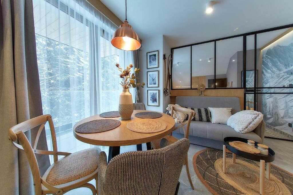 A modern living room with a round wooden table, wicker chairs, a sofa, and a large window with a view of trees.