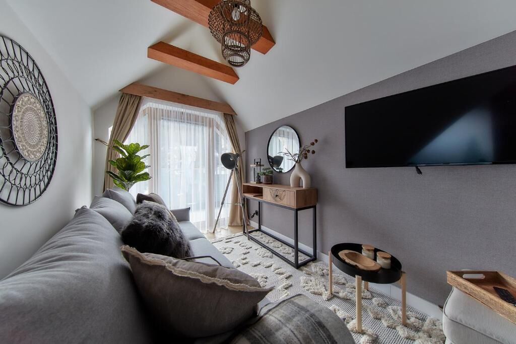 Un moderno salón con un sofá gris, una mesa de centro de madera, un televisor montado en la pared y un gran ventanal con cortinas.