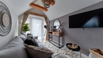Un moderno salón con un sofá gris, una mesa de centro de madera, un televisor montado en la pared y un gran ventanal con cortinas.
