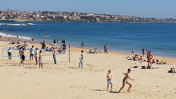 Una spiaggia con persone che giocano a pallavolo, una riva sabbiosa e una città sullo sfondo.