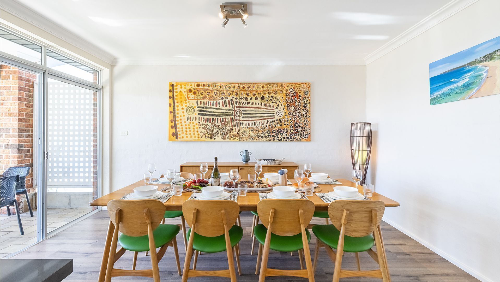 A dining area with a large table set for six, surrounded by wooden chairs with green cushions. A large abstract painting hangs above the table, and a beach scene painting is on the wall to the right.