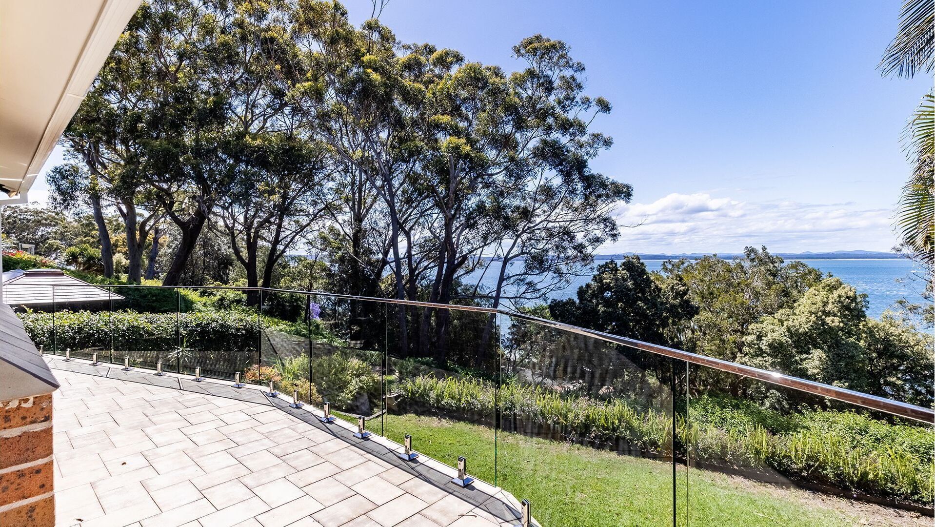 A balcony with a glass railing overlooking a lush garden and a coastal view.