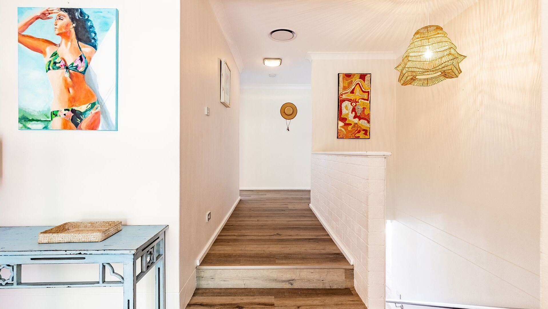 A hallway with wooden flooring, a staircase, a wall-mounted light fixture, and two framed artworks.