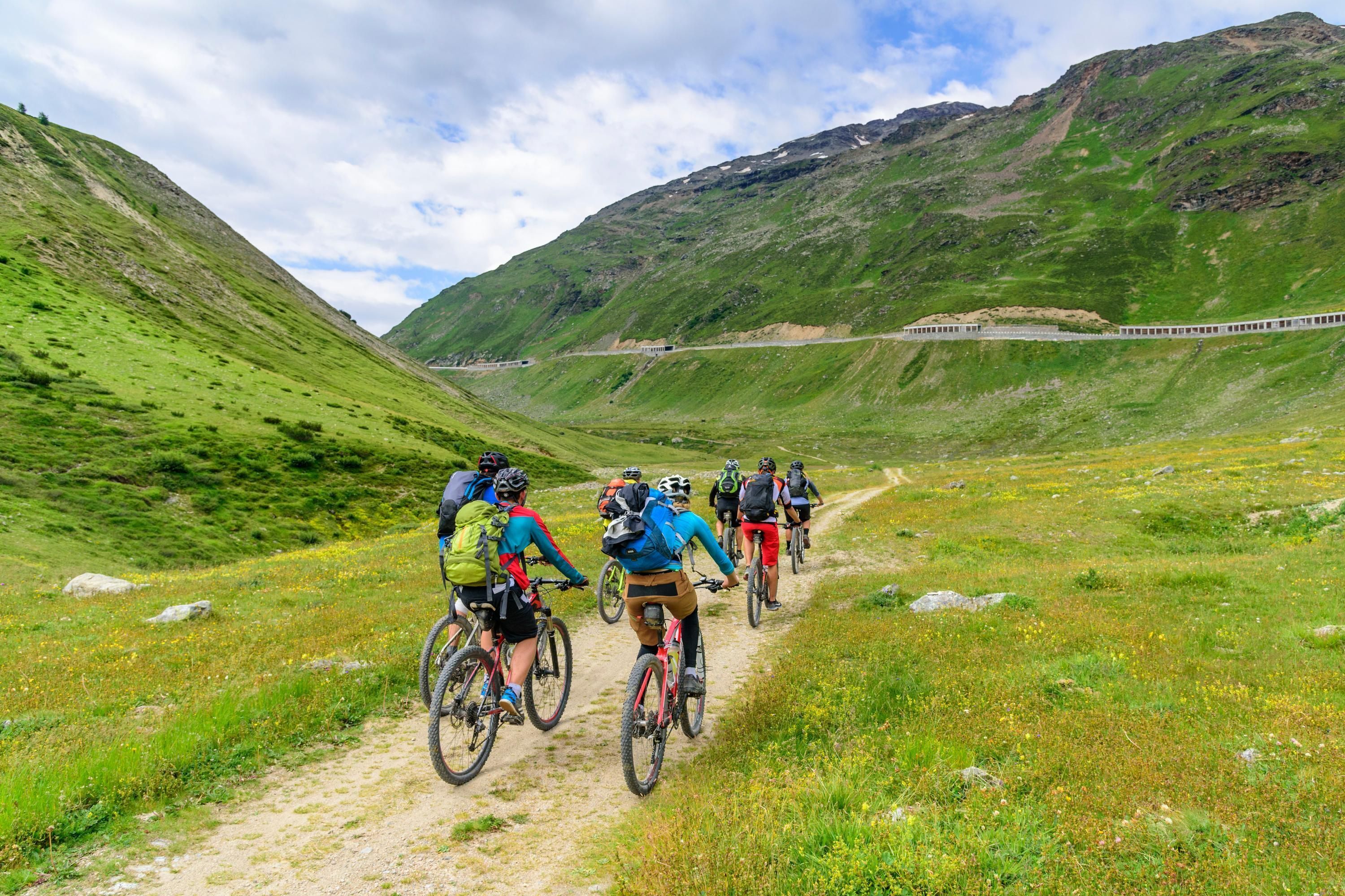 A group of cyclists on a dirt path in a mountainous area with green hills and a clear sky.