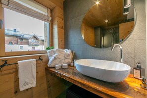 A modern bathroom with a wooden interior, a large oval sink, a round mirror, and a window offering a view of a building outside. - Lina in Livigno With 2 Bedrooms and 2 Bathrooms (Livigno)