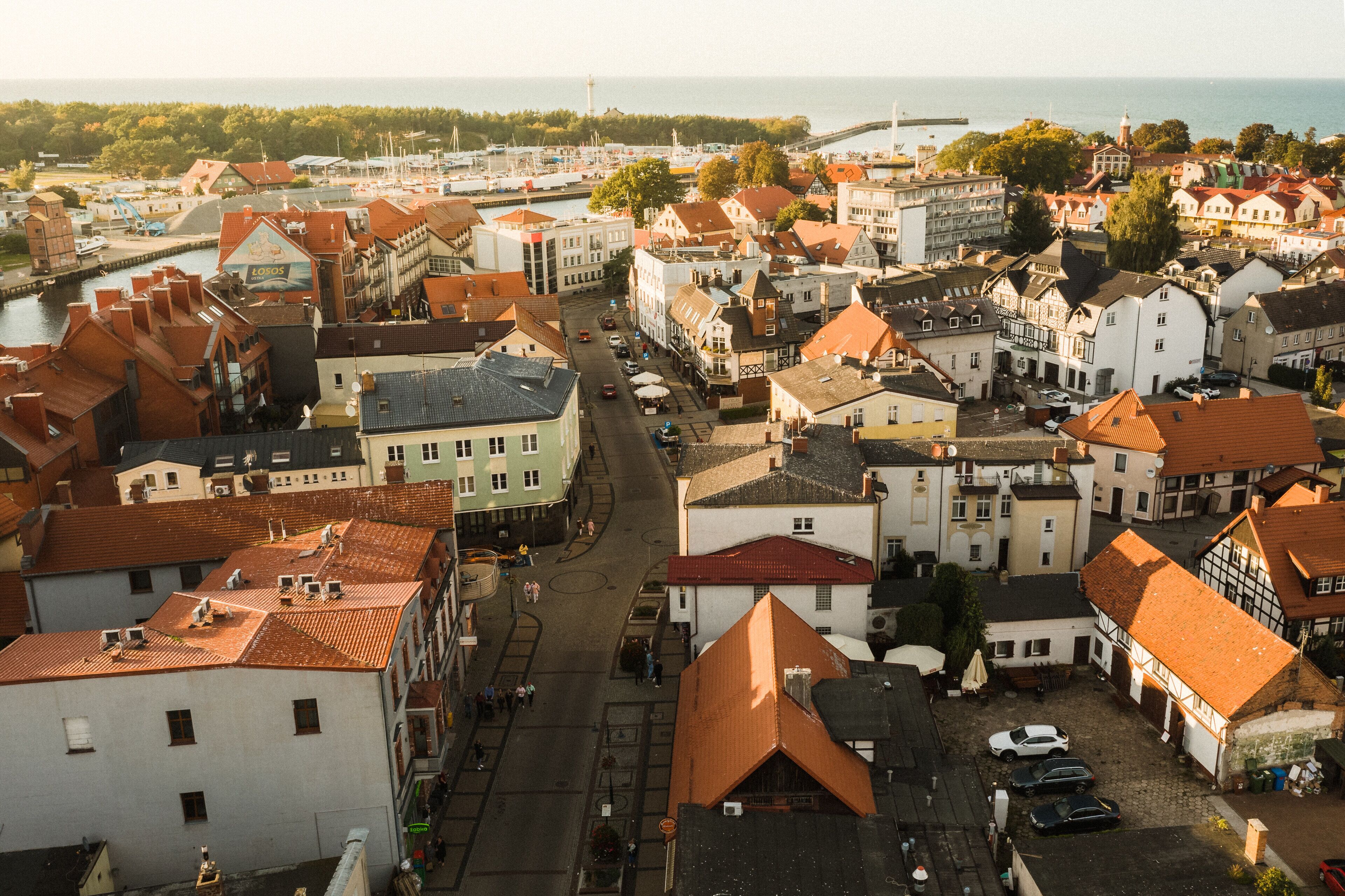 A town with a central street lined with historic buildings, a marina in the background, and a view of the sea.