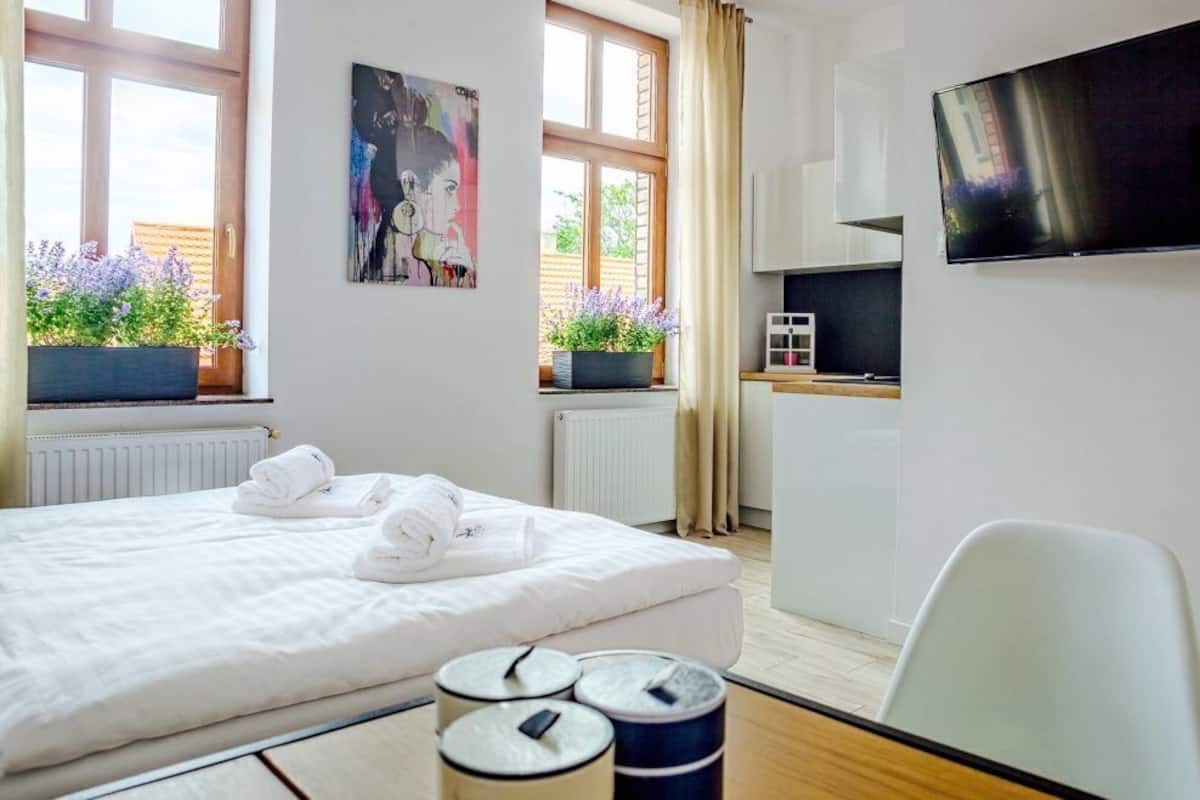 A hotel room with a bed, a TV, a desk, and a window with plants.
