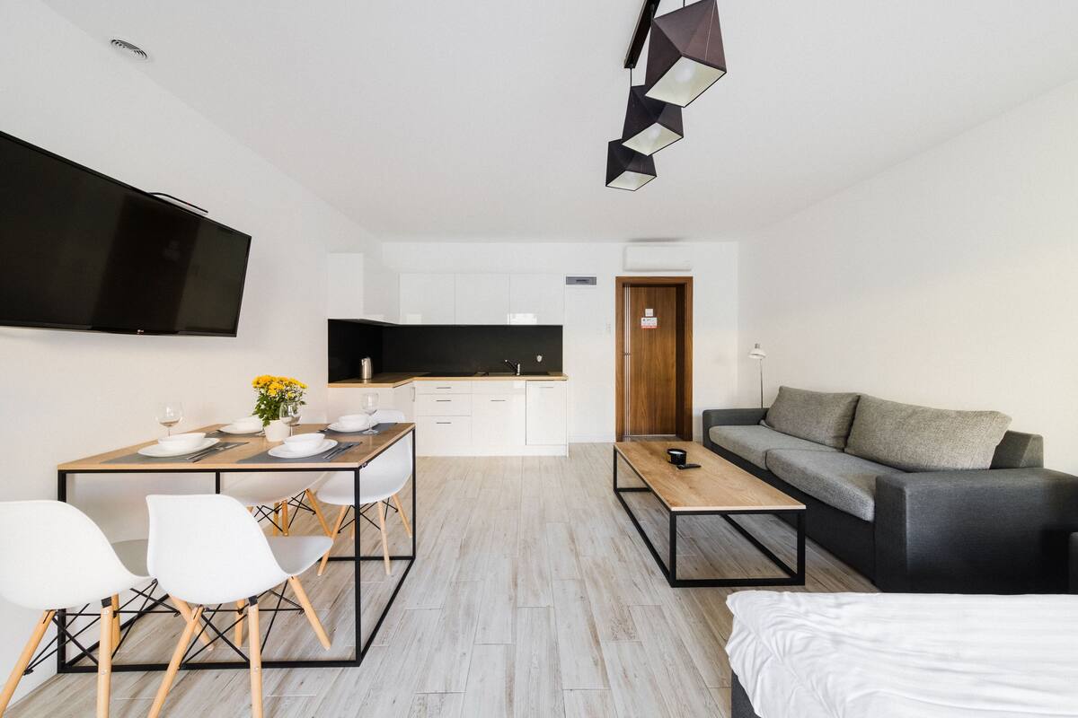 A modern living space with a dining area, a sofa, a TV mounted on the wall, and a kitchen in the background.