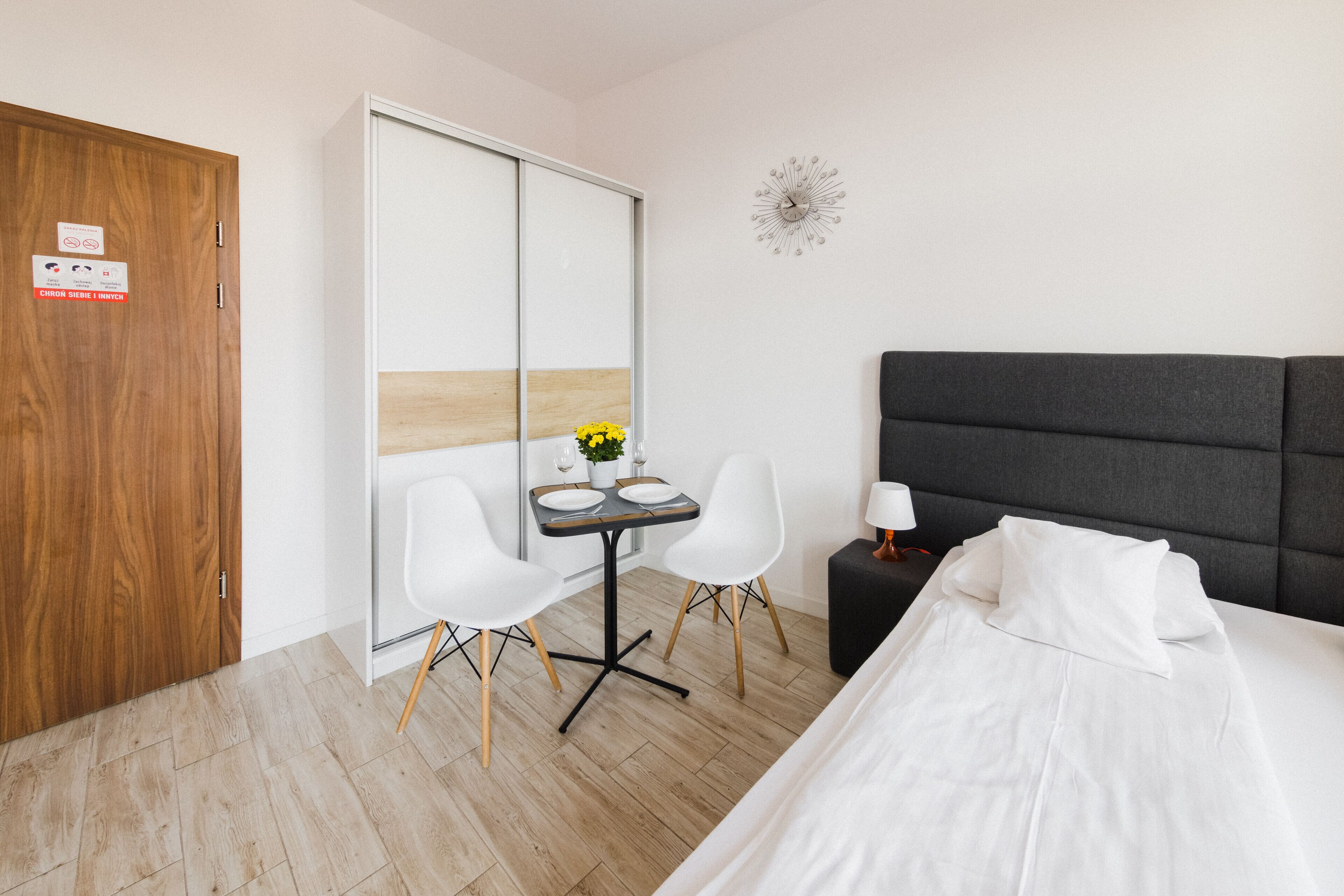 A hotel room with a bed, a desk with two chairs, a small table with a vase of flowers, and a clock on the wall.