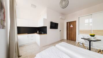 Quarto de hotel moderno com uma cama, uma kitchenette compacta, uma área de jantar com mesa e cadeiras, e uma TV fixada na parede.