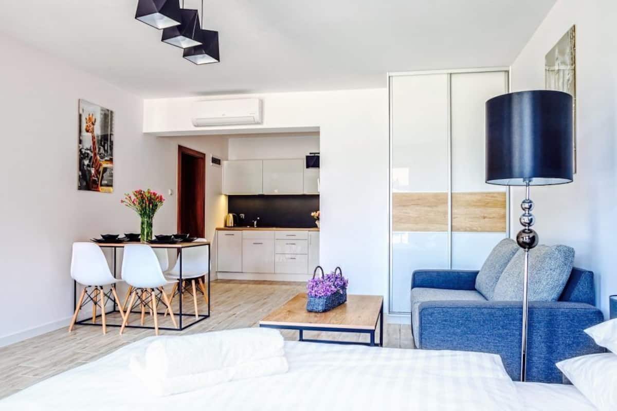 A modern hotel room with a bed, a sofa, a dining table with chairs, a kitchen area, and a large window.