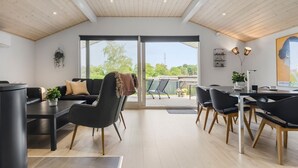 A modern living room with a black sofa, a dining table, and chairs. There is a large window providing a view of the outdoors. - 3 bedroom stunning home in Hejls (Hejls)