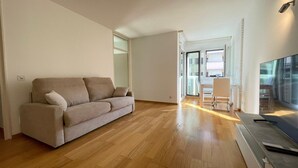 A spacious living room with a beige sofa, wooden flooring, and a large flat-screen TV.