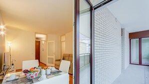 A modern living room with a glass door leading to a balcony, a dining area with a table set for two, and a cozy seating area with a sofa and armchair.