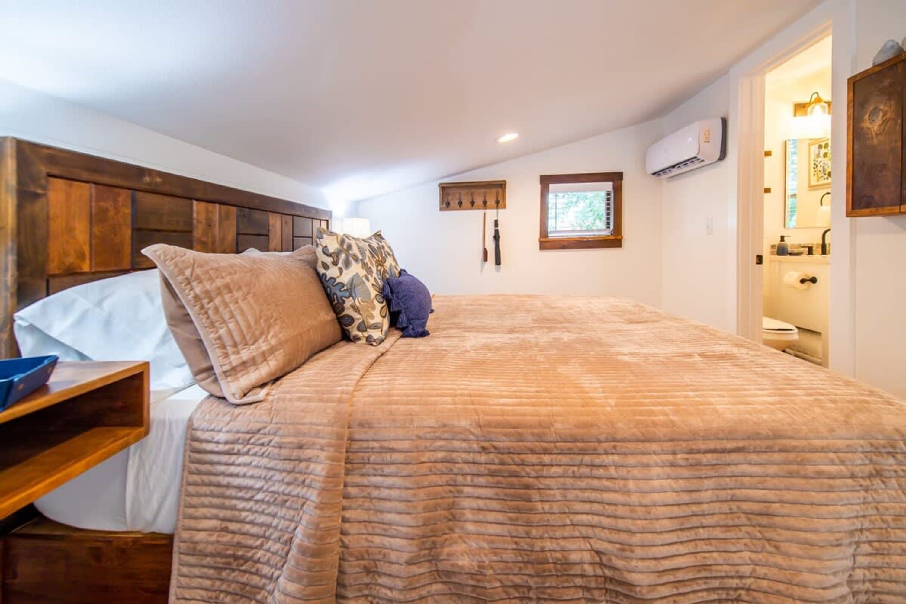 A bedroom with a wooden headboard, a large bed with a textured blanket, a bedside table with a lamp, and a small window with a view.