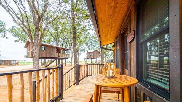 Eine Holzterrasse mit rundem Tisch und Bänken, umgeben von Bäumen und Holzhütten.