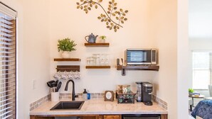 Cocina compacta con lavamanos, microondas y cafetera. Hay armarios de madera y una pequeña planta en un estante.