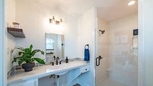 Un baño moderno con tocador blanco, espejo, planta y ducha con mampara de vidrio.