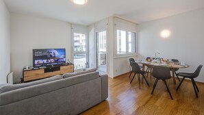 Un salon moderne avec un canapé gris, une table à manger en bois avec des chaises, un téléviseur posé sur un meuble en bois et de grandes fenêtres laissant entrer la lumière naturelle.