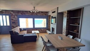 A modern living room with a wooden floor, a sofa, a dining table with chairs, a TV, and a fireplace. - Maritinas Stone Apartment On The Beach (Pyrgos)