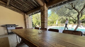 Un espace extérieur abrité, aménagé pour les repas, avec une longue table en bois, des chaises et un barbecue.