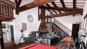 Une table de billard se trouve dans une pièce rustique, avec des poutres en bois apparentes, une cheminée en pierre et un escalier menant à un autre niveau.