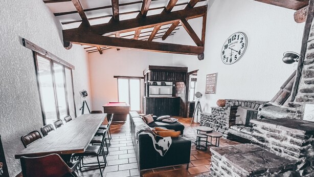 Une pièce spacieuse avec une longue table en bois et des chaises, un coin salon confortable avec un canapé et un pouf, une cheminée en pierre et une horloge au mur.