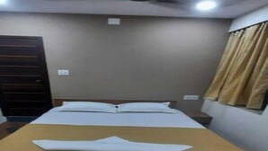 Una cama individual con una colcha marrón en una habitación con suelo de madera, ventilador de techo y una ventana con cortinas.