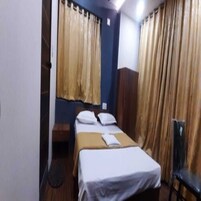 Una habitación pequeña con cama individual, cabecera de madera, mesita de noche y dos cortinas.