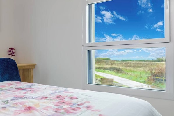 A room with a bed, a chair, a wooden table, and a window with a view of a rural landscape.