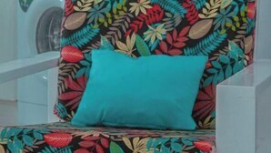 Une chaise ornée d’un motif de feuilles aux couleurs vives et dotée d’un coussin turquoise, dans une pièce dont un mur est recouvert de carreaux et où l’on aperçoit une machine à laver à l’arrière-plan.