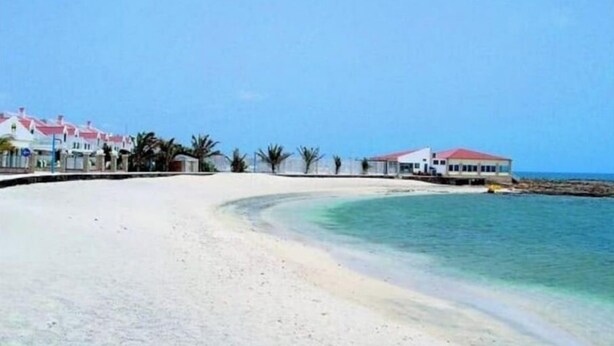 Une plage de sable blanc bordée d’une eau turquoise et limpide. Plusieurs bâtiments aux toits rouges se dressent le long du littoral.