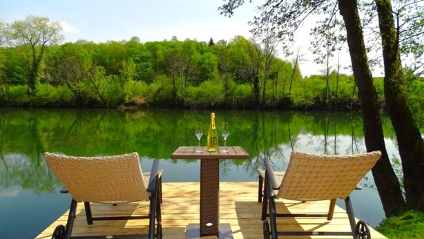 A wooden deck with two wicker chairs and a table, overlooking a calm river. - House Kolpa Paradise Whirlpool And Sauna (Metlika)