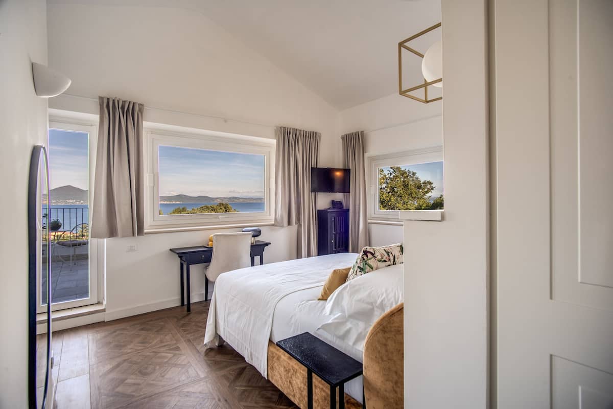 Ein Schlafzimmer mit Bett, Schreibtisch und Stuhl, großen Fenstern mit Vorhängen und Blick ins Freie.