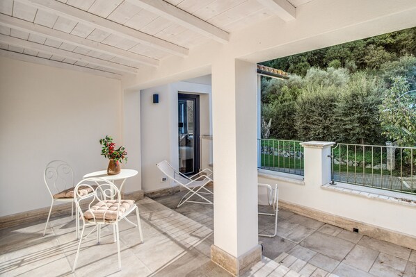 A balcony with a white chair, a small table with a vase, and a view of greenery outside.