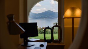 Un escritorio con monitor de computadora, teclado y ratón, situado cerca de una ventana que ofrece vistas a un lago y montañas.