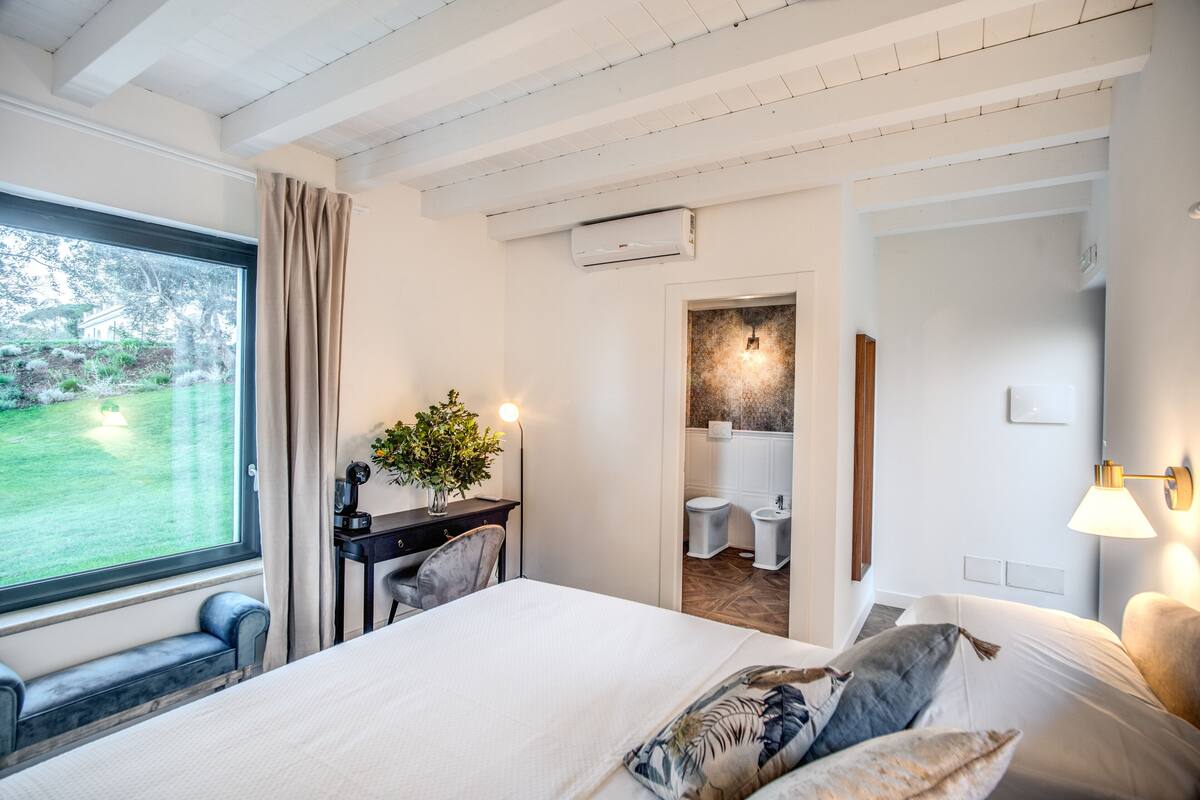 Ein Schlafzimmer mit einem großen Bett, einem Schreibtisch mit Pflanze, einem Fenster mit Vorhängen und einer Klimaanlage.