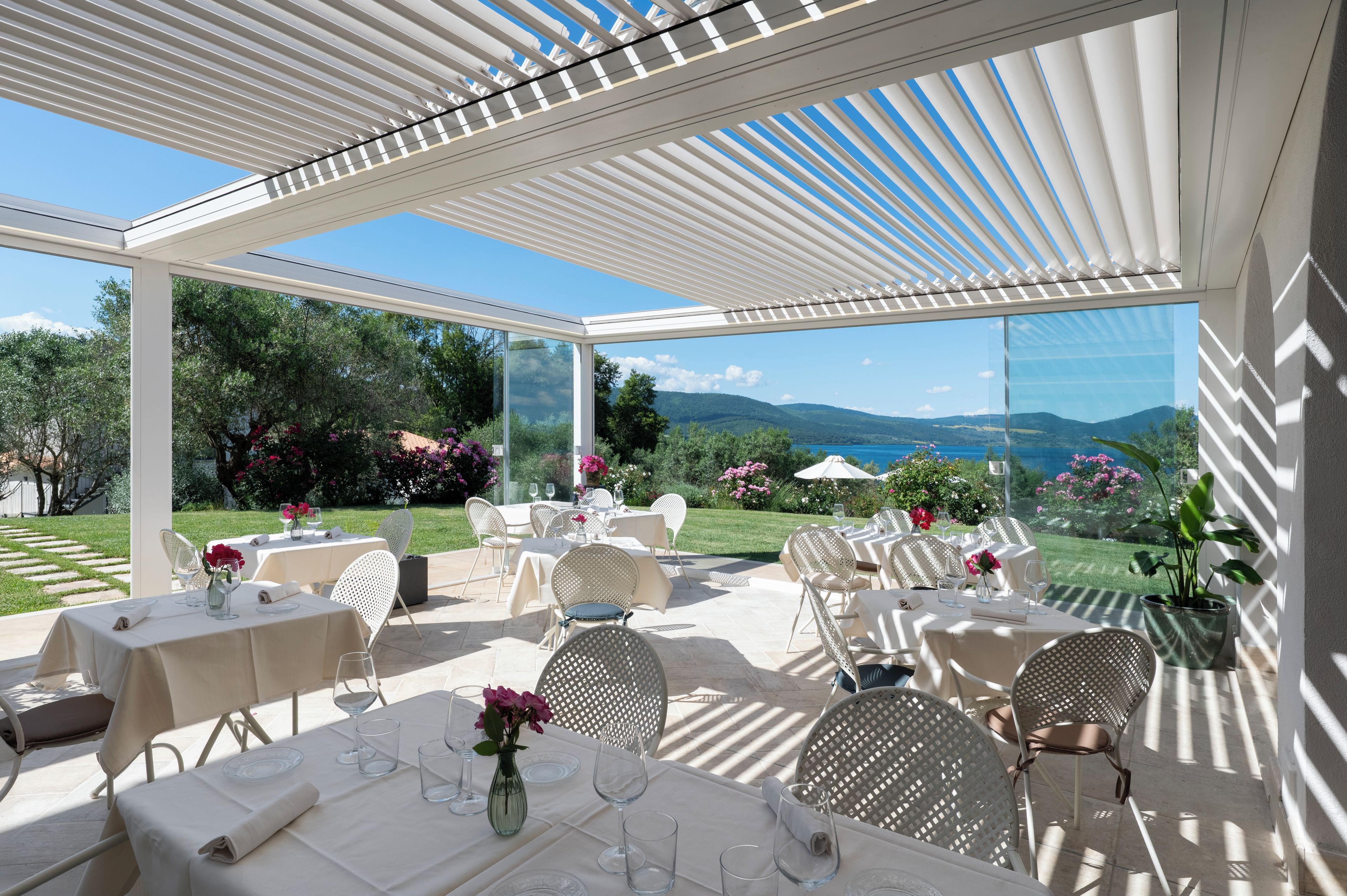 An outdoor dining area with tables and chairs, a pergola overhead, and a scenic view of hills and a body of water.