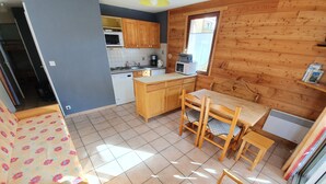A compact kitchen with wooden cabinets, a microwave, and a coffee maker. There is a dining area with a table and chairs, and a sofa with a floral pattern. - 002 Rochebrune - Chalet l'Hysope (La Salle-les-Alpes)