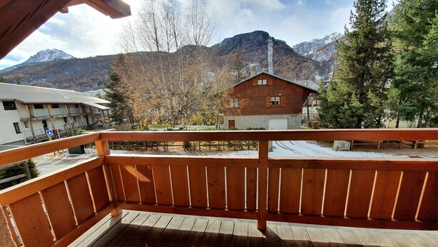 A wooden balcony with a view of a mountain and a building with a red roof. - 002 Rochebrune - Chalet l'Hysope (La Salle-les-Alpes)