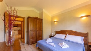 A bedroom with a bed, a nightstand, a lamp, a wooden wardrobe, and a vase with branches. - One bedroom apartment (4 people) Premium - Residence Baia delle Palme (Sardegna)