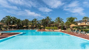 A large outdoor swimming pool with multiple lanes, surrounded by palm trees and shaded areas with umbrellas. - One bedroom apartment (4 people) Premium - Residence Baia delle Palme (Sardegna)